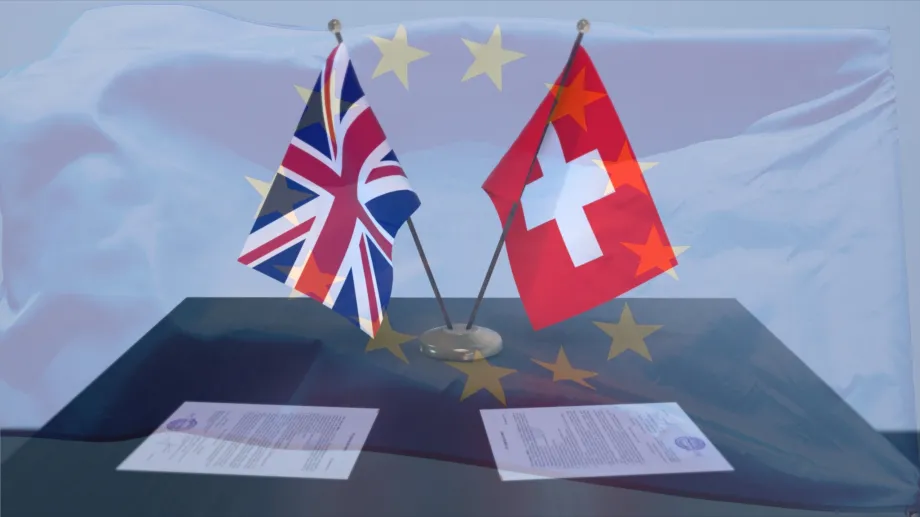 Na stolu dvije male zastave Velike Britanije i Švicarske, a ispred njih dva službena dokumenta. U pozadini se nazire zastava Europske unije.