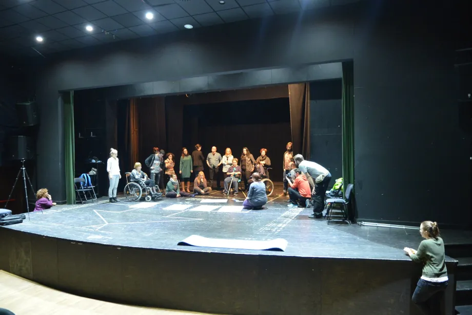 Grupa ljudi stoji i sjedi na pozornici. Pored pozornice su dvije žene, svaka s jedne strane