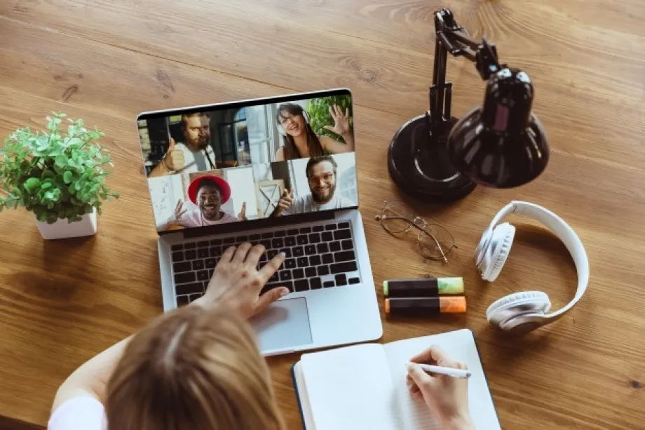 Žena sjedi za stolom, u jednoj ruci drži kemijsku, drugu ruku drži na tipkovnici laptopa na kojemu je prikaz online sastanka. pored laptopa su na stolu bilježnica, biljka, svjetiljka, slušalice i naočale