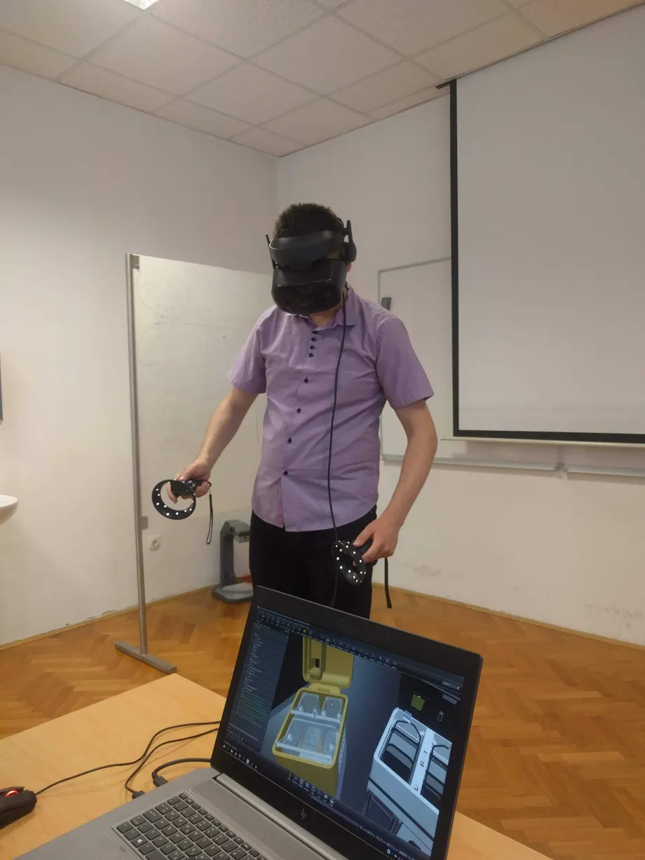Muškarac stoji u dvorani, na glavi i u rukama drži modele proizvoda virtualne stvarnosti