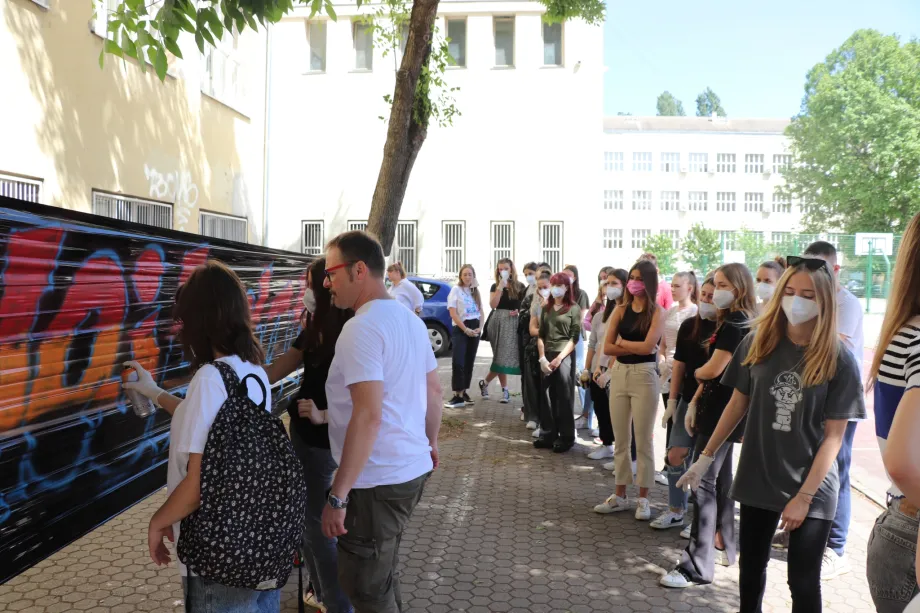 Dvije učenice izrađuju grafit, između njih stoji muškarac, a iza njih stoji grupa učenika koji gledaju izradu grafita