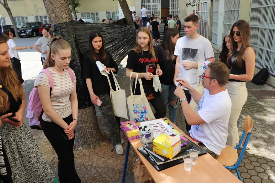 Muškarac sjedi za stolom i priča, a grupa učenika stoji ispred i pored stola i slušaju