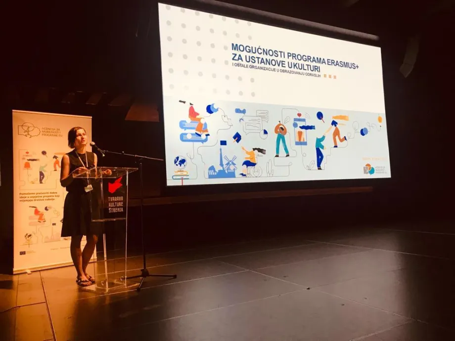 Govornica drži govor na pozornici ispred prezentacijskog platna na kojemu piše naslov "Mogućnosti programa Erasmus+ za ustanove u kulturi i ostale oranizacije u obrazovanju odraslih"