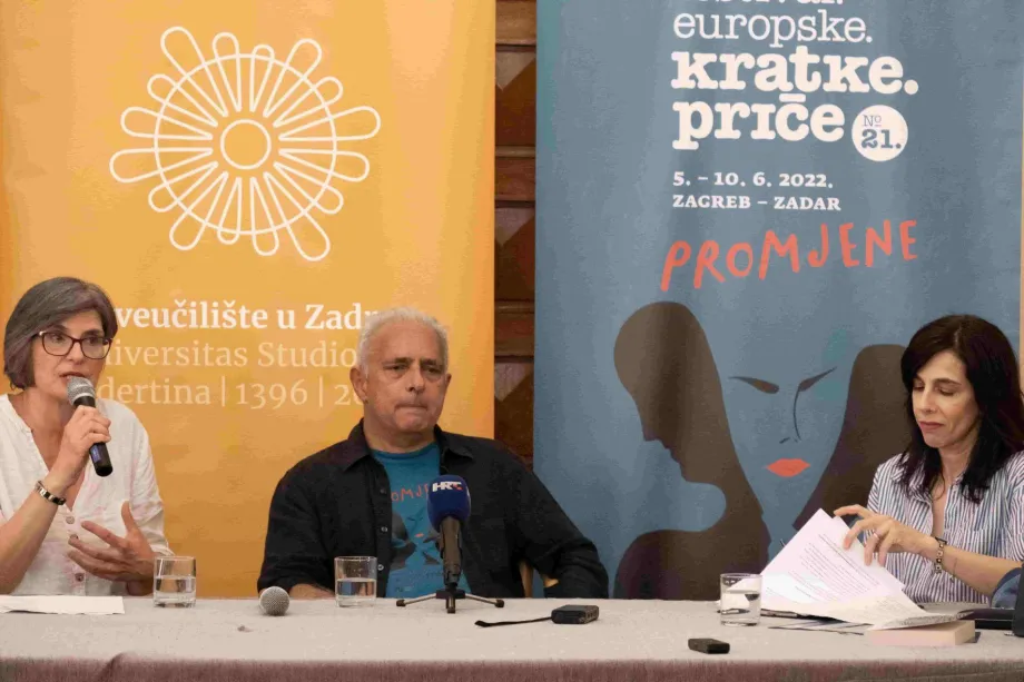 Dvije žene i jedan muškarac sjede za stolom, jedna žena drži mirofon u ruci i priča, druga žena lista papire