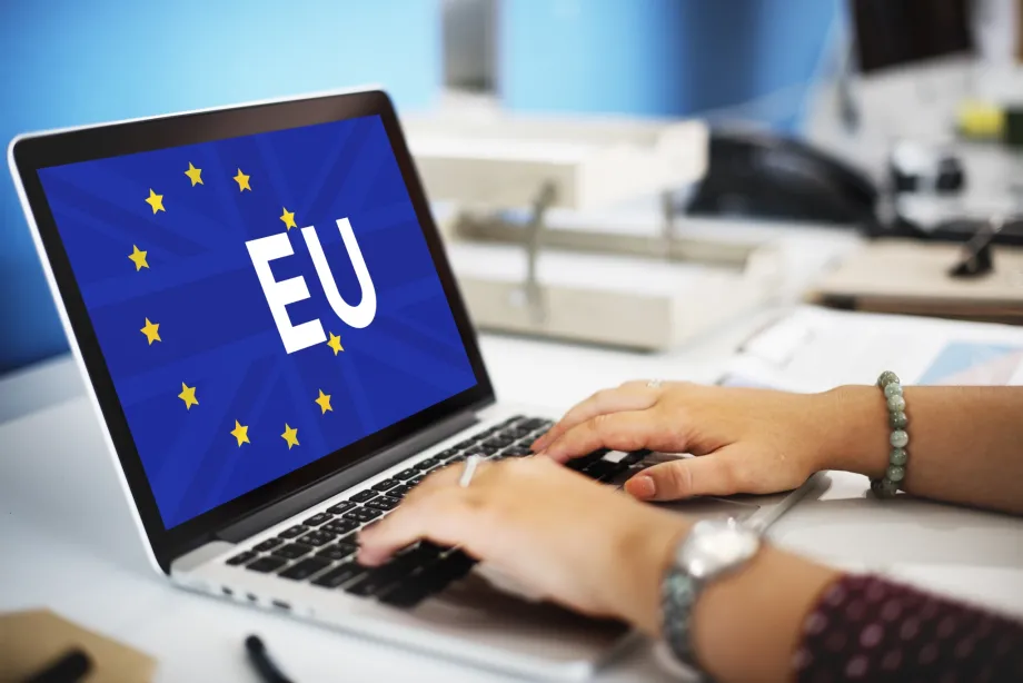 Ruke koje tipkaju po laptopu na čijoj je pozadini vidljiva zastava Europske unije i natpis EU