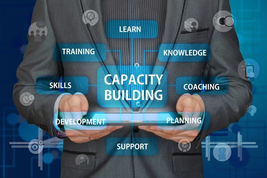 Muškarac u odijelu drži u rukama ispred sebe tablet iz kojega se kao hologram prikazuje pravokutnik u kojem piše "capacity building" i s njim su spojeni drugi pravokutnici u kojima piše skills, training, learn, knowledge, coaching, development, support, planning.