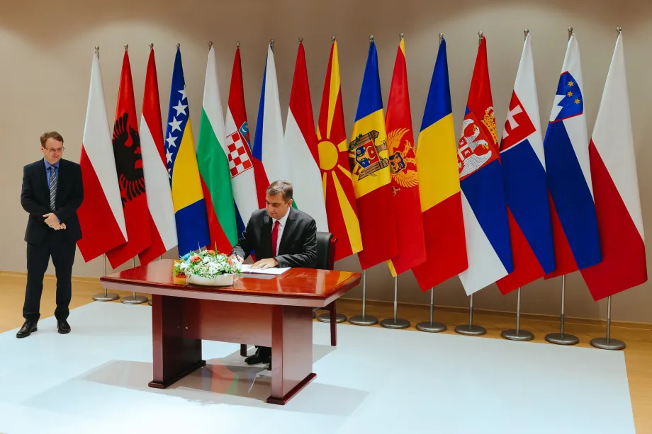 Muškarac sjedi za stolom i potpisuje ugovor, drugi muškarac stoji malo dalje od stola. Iza njih nalaze se zastave europskih država.