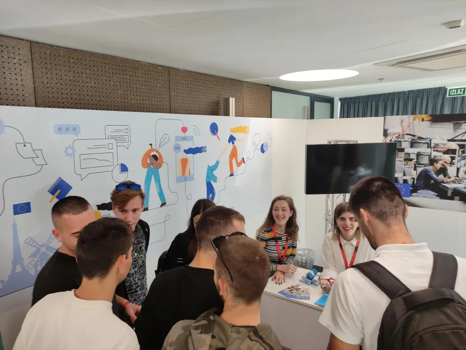Mladići stoje kod štanda AMPEU-a na Expo sajmu dok im dvije djevojke objašnjavaju o programima EU.