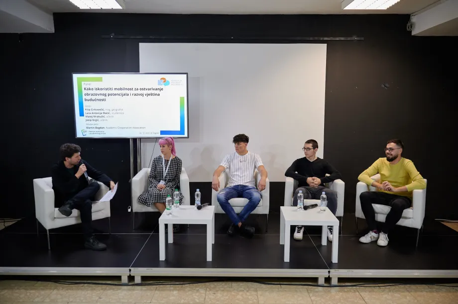 Moderator panela govori na mikrofon dok ga četvero mladih panelista sluša. Sjede u stolcima na pozornici, a iza njih je natpis "Kako iskoristiti mobilnost za ostvarivanje obrazovnog potencijala i razvoj vještina budućnosti."
