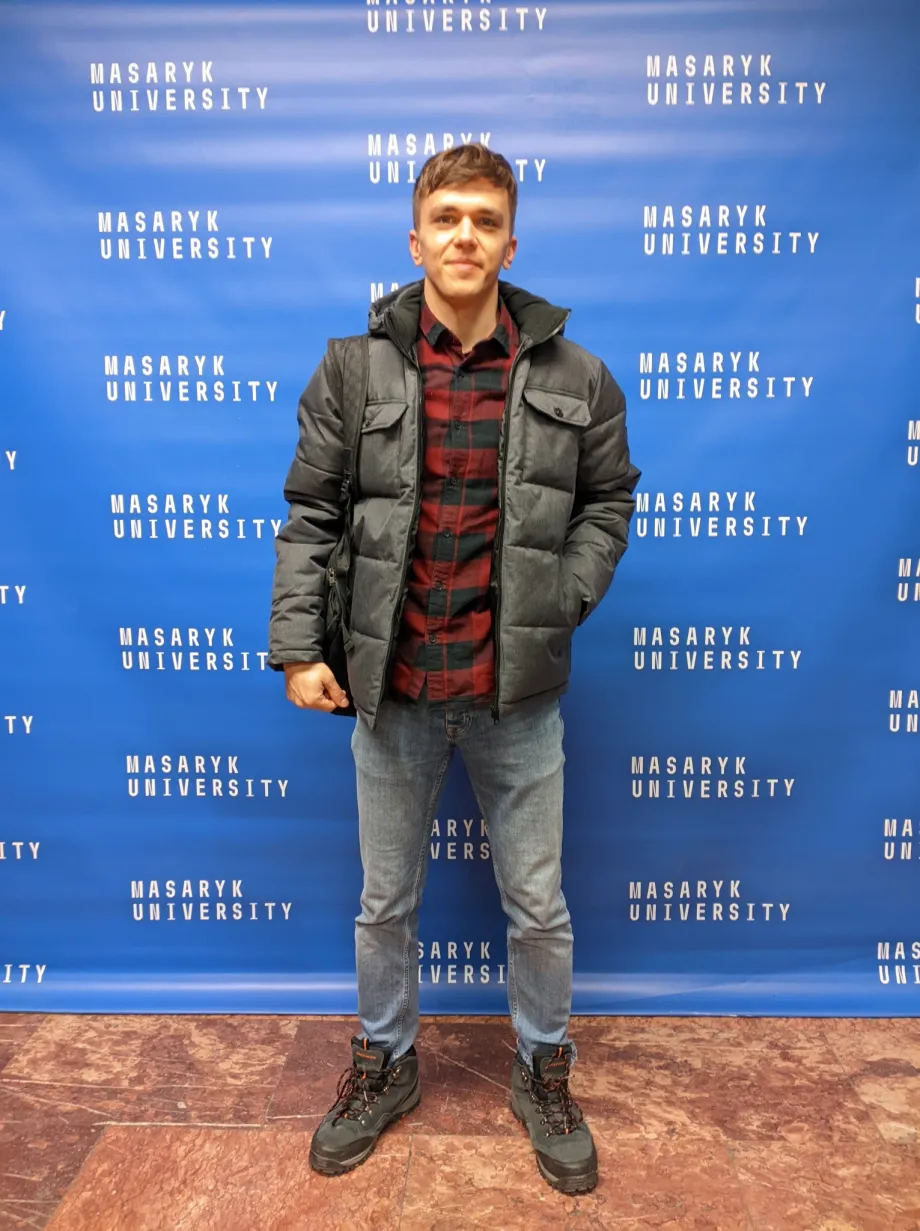 Filip Fila stoji, iza njega je plavi zid na kojem piše mnogo puta "Masaryk University".