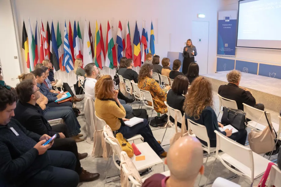 Žena drži predavanje pred punom dvoranom. Na zidu uz publiku nalaze se zastave država članica EU.