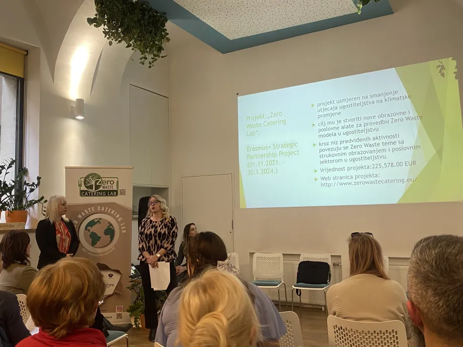 Grupa polaznika okrenutih leđima i dvije žene koje stoje ispred njih i drže prezentaciju. Na zidu je otvorena prezentacija na kojoj piše " Projekt Zero Waste Catering Lab"