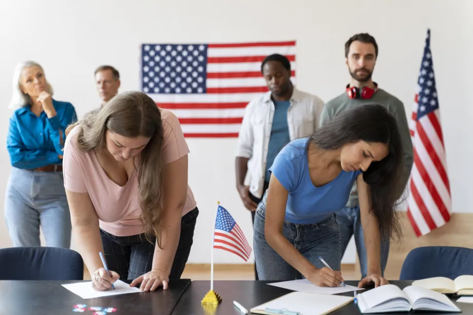 Dvije ženske osobe upisuju podatke u zadaće. Na fotografiji je ukupno šestero ljudi različitog spola i rase u zajedničkoj prostoriji ukrašenoj američkim zastavama