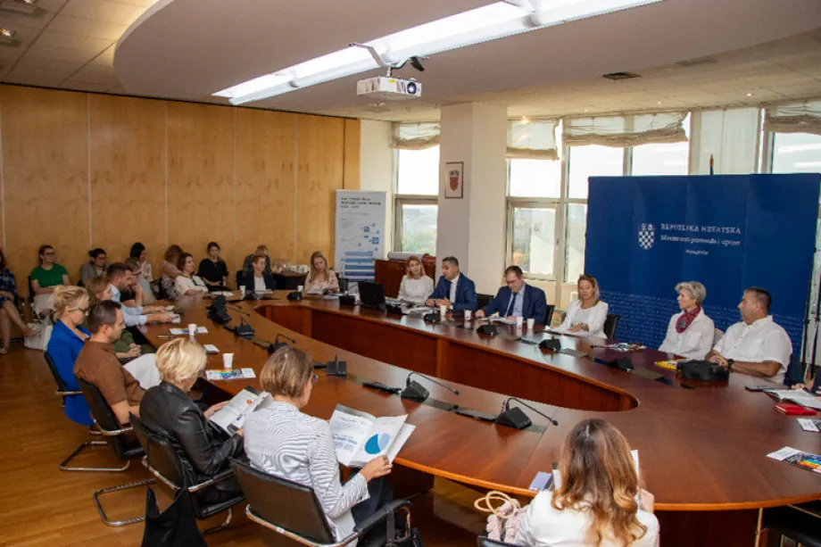 Okrugli stol s oko 20-tak osoba, u pozadini veliki reklamni zid sa službenim logotipom Miinistarstva pravosuđa, uprave i digitalne transformacije