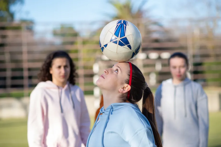 Tri djevojke na sportskom terenu, od kojih jedna vješto balansira loptu glavom, prikazuju timsku igru, vježbu i zdrav način života u sportskoj odjeći, pokazujući svoje vještine i posvećenost treniranju.