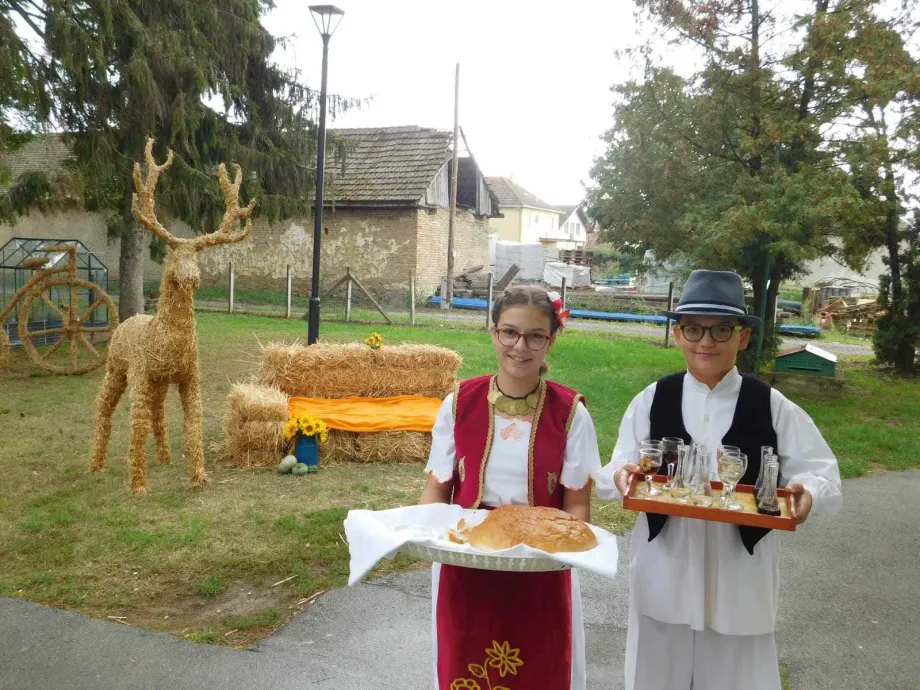 Djevojčica u narodnoj nošnji nosi pladanj s kruhom, a dječak nosi pladanj s pićem, u pozadini je seosko dvorište sa slamnatom figurom jelena i slamnatim kaučom