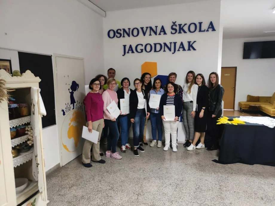 13 učitelja pozira ispod natpisa na zidu Osnovna škola Jagodnjak, u prednjem lijevom kutu je drveni ormar u kojem se nalaze teglice sa zimnicom i zdjelam a u gornjem desnom kutu je stol na kojem se nalazi cvijet suncokreta