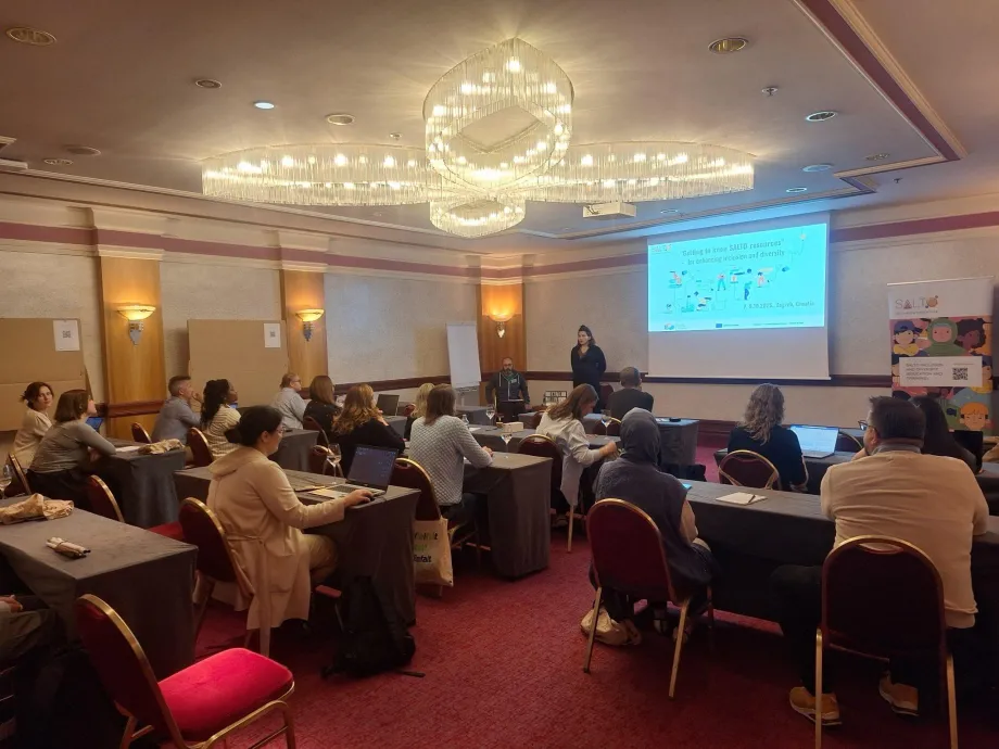 Hotelska dvorana u školskom postavu u kojoj se nalaze odrasli polaznici u klupama i slušaju pozdravni govor ženske osobe koja stoji na sredini dvorane, a pored nje je projektno platno na kojem se nalazi prvi slajd prezentacije