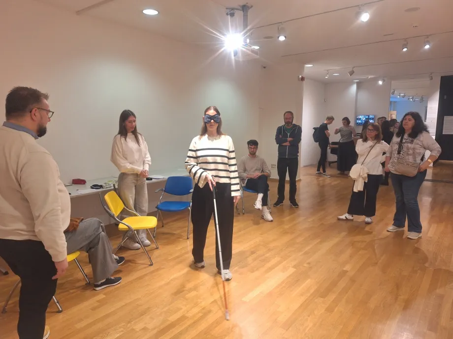 Ženska osoba s povezom preko očiju pokušava se kretati uz pomoć bijelog štapa za slijepe i slabovidne, a šest osoba oko nje ju promatra. U donjem dijelu dvorane vide se osobe koje međusobno razgovaraju.