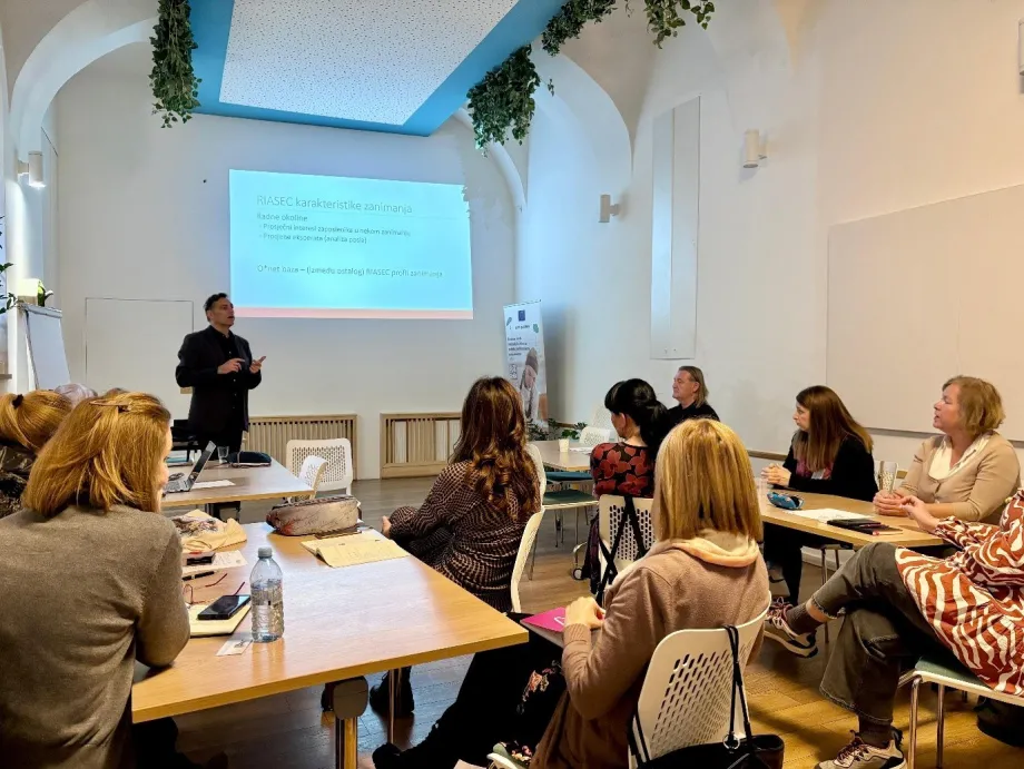 Skupina polaznika u školskim klupama i muškarac ispred projekcijskog platna drži predavanje