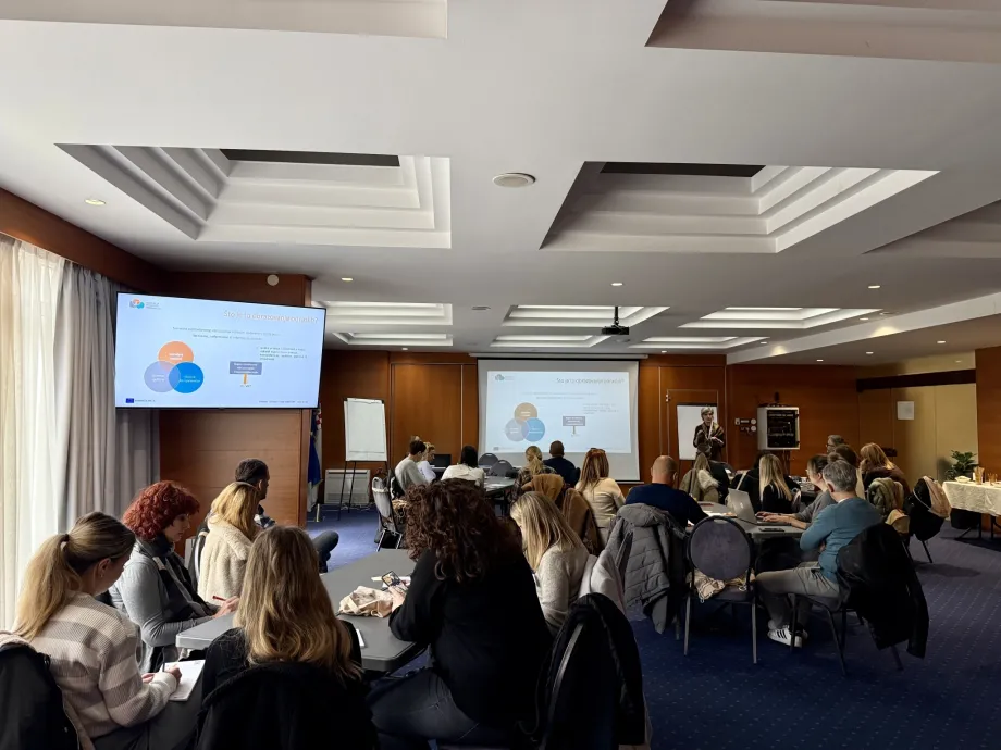 Predavaonica sa sudionicima podijeljenim po skupinama, a ekranima se vidi prezentacija