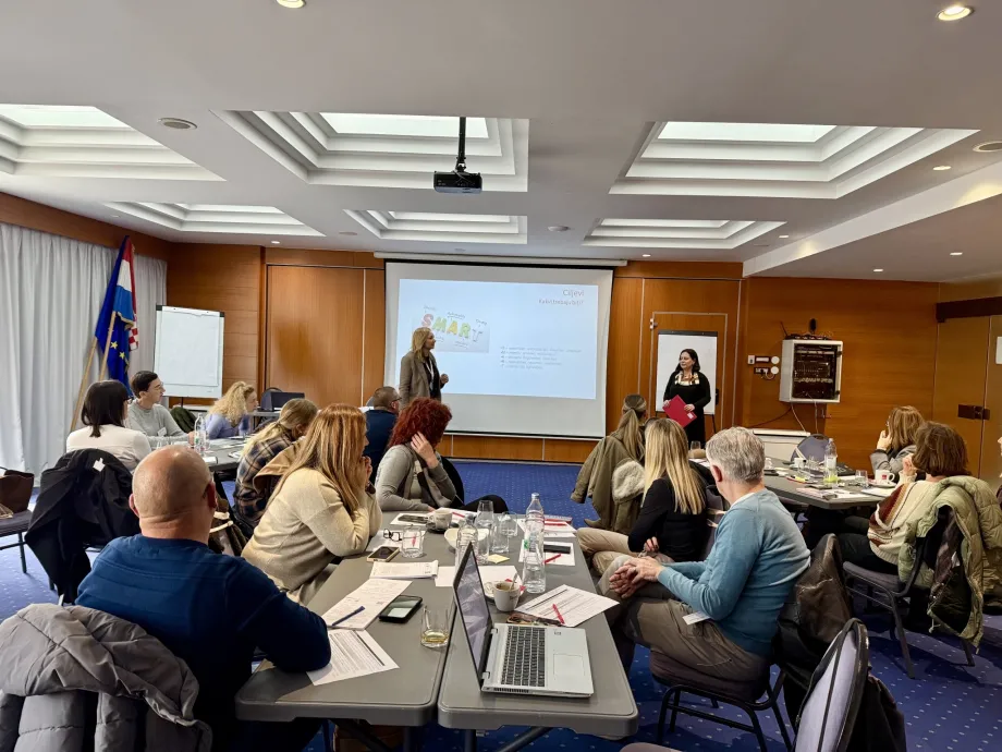 Predavaonica sa sudionicima podijeljenim po skupinama. Neki od njih imaju laptope na stolu, a ekranima se vidi prezentacija