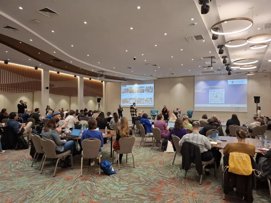 Kongresna dvorana u kojoj sudionici sjede za okruglim stolovima i prate izlaganja na dva projekcijska platna