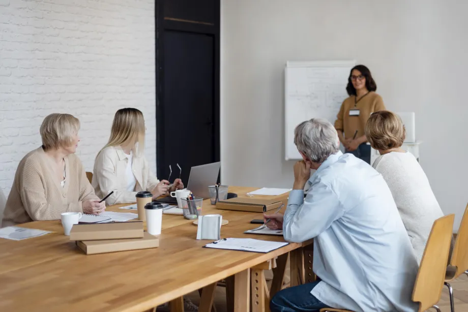 Poslovni sastanak u uredu gdje za stolom sjede četiri osobe dok peta predaje ispred flip-charta