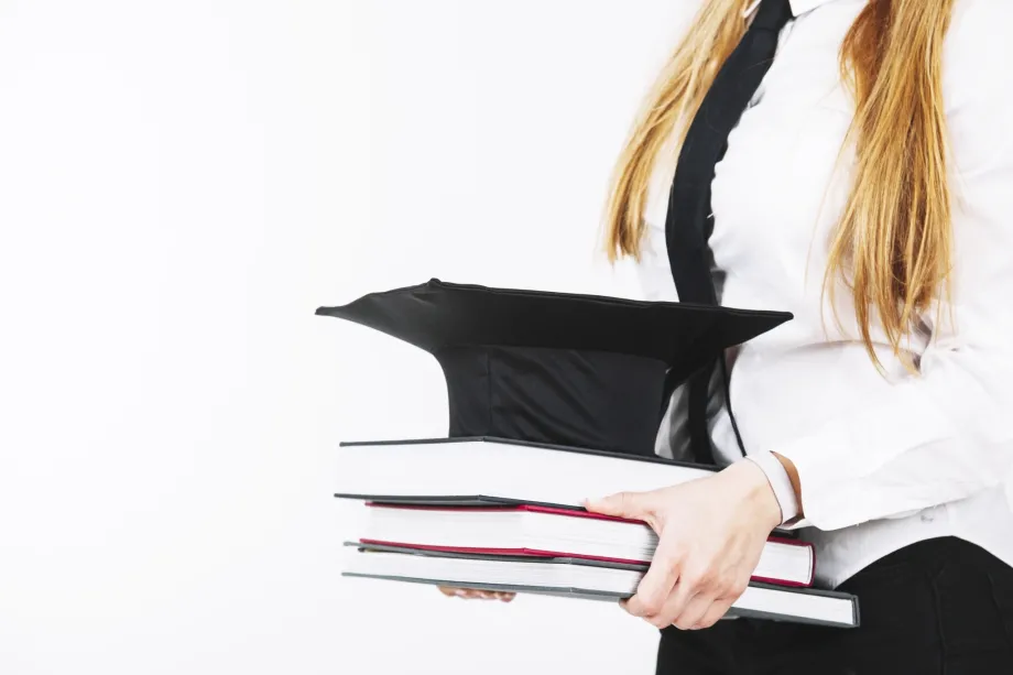 Ženska osoba plave kose kojoj se ne vidi glava ispred sebe nosi tri knjige s akademskom kapom