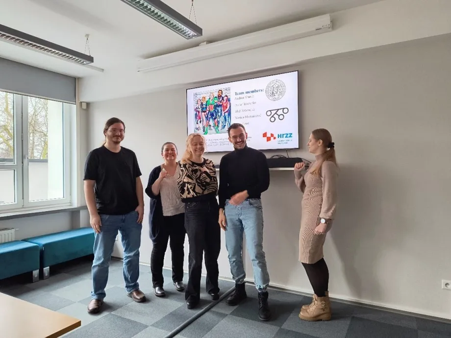 Petero nasmiješenih osoba, tri žene i dvije muškarca, stoje nasmiješeni ispred ekrana